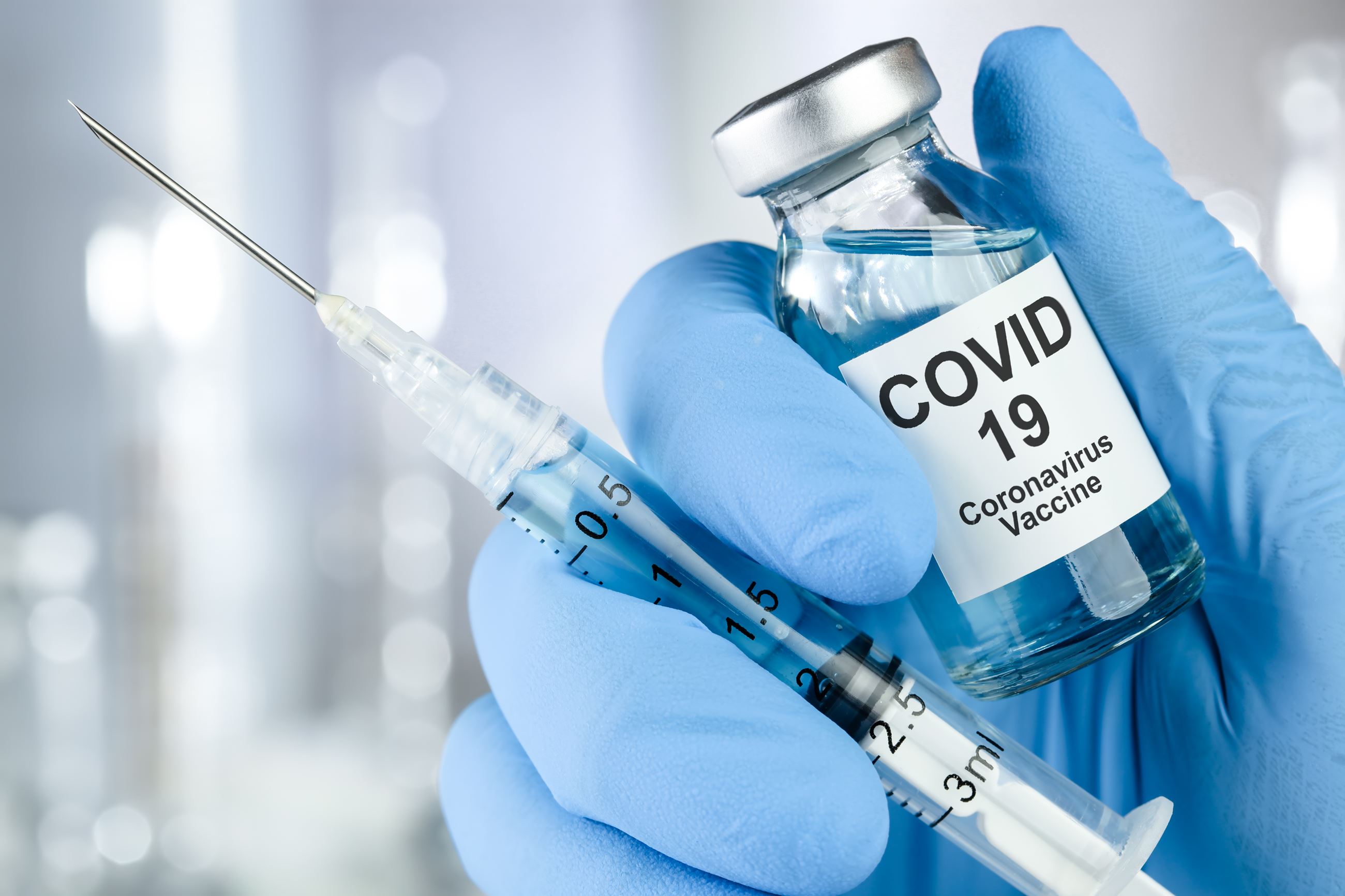 Hand in blue-glove holding COVID-19 vaccine vial and needle.